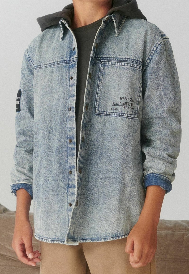 Personne portant une veste en jean bleu clair délavée sur un sweat à capuche foncé, avec les manches retroussées et un pantalon beige, debout à l'intérieur.