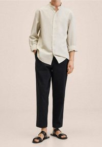 Camicia button-up beige chiaro con colletto alla coreana, pantaloni scuri e sandali neri. Vestibilità casual con maniche arrotolate e texture del tessuto liscia.