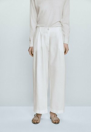Personne portant un pantalon blanc taille haute à jambes larges, un haut beige à manches longues et des sandales plates à brides, debout devant un fond clair uni.