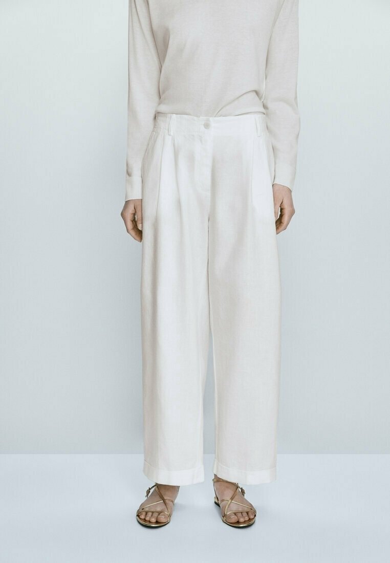Personne portant un pantalon blanc taille haute à jambes larges, un haut beige à manches longues et des sandales plates à brides, debout devant un fond clair uni.