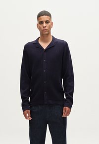 Marinblå stickad cardigan med slagkrage, knäppning fram med knappar och textilstruktur. Buren över mörka byxor, med långa ärmar.