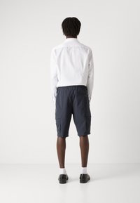 Chemise blanche à manches longues, short cargo bleu marine, chaussures noires. Le short comporte deux poches latérales et une taille élastique. Fond uni.