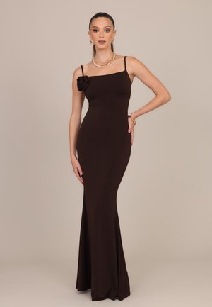 Femme portant une longue robe noire ajustée avec fines bretelles et un détail floral sur une épaule, accessoirisée avec des bijoux en or.