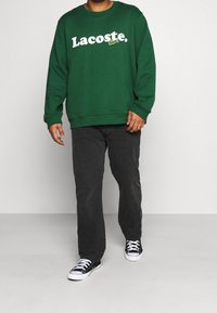 Felpa verde in cotone con logo "Lacoste" bianco e accento di coccodrillo, abbinata a jeans scuri e sneakers nere.