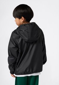 Bambino con capelli neri corti che indossa una giacca nera con cappuccio, una maglietta bianca e pantaloni verdi, rivolto di lato su uno sfondo semplice.