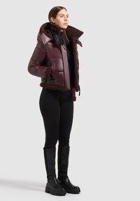 Femme portant une doudoune bordeaux foncé brillante avec une doublure en peau retournée marron, des leggings noirs et des bottes noires hautes jusqu'aux genoux, debout sur un fond uni.