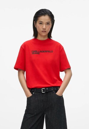 Femme aux cheveux raides et foncés porte un t-shirt rouge Karl Lagerfeld Jeans et un pantalon noir à fines rayures avec une ceinture noire, debout les mains dans les poches.