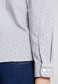 Camicia a maniche lunghe di colore grigio chiaro con sottili strisce verticali e piccoli punti blu navy. Presenta polsini abbottonati e un orlo curvo.
