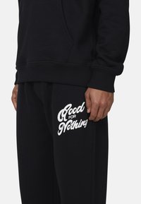 Zwarte hoodie met kangoeroezak, gecombineerd met zwarte joggingsbroek met witte tekst "Good for Nothing" op de bovenbeen.