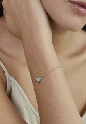 Silver chain bracelet featuring a heart charm with a glossy white inlay, worn on the wrist. Smooth texture, delicate design.