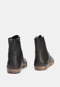 Botas de tobillo de cuero negro con un upper texturizado negro, cremallera lateral y suela de goma marrón claro con un ligero patrón de relieve.