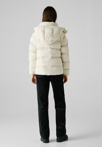 Puffjacke in Cremefarbe mit horizontalen Steppnähten und Kapuze, kombiniert mit schwarzen Cordhosen und kräftigen schwarzen Stiefeln.