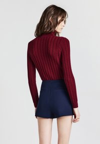 Bordeaux gestrickter Pullover mit Rippmuster, hohem Kragen und langen Ärmeln, kombiniert mit navyblauen, taillenhohen Shorts mit Gesäßtaschen.