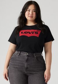 Tricou negru cu un logo roșu Levi's pe piept, asortat cu blugi gri închis, cu talie înaltă. Materialul este moale; designul este casual.