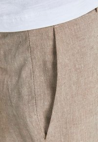 Pantalones en lin beige avec une texture lisse, des détails de plis et des poches latérales. Le matériau semble léger et respirant.