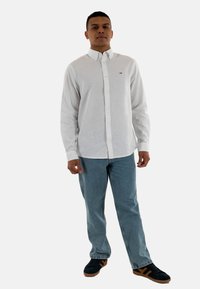 Camicia bianca a maniche lunghe con bottoni in cotone, abbinata a un paio di jeans in denim blu chiaro e sneakers nere con dettagli marroni.