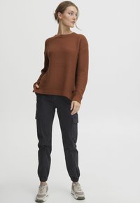 Pull en tricot texturé marron avec col rond, associé à un pantalon cargo noir avec poches latérales et poignets élastiques, complété par des baskets.