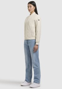 Ljust beige quiltad bomberjacka med ståkrage, ribbade ärmar och en rund logotypaccent; kombinerad med ljusblå raka jeans och vita sneakers.
