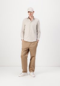 Stripet skjorte i beige og hvit, brune bukser og hvite sneakers. Tilbehøret inkluderer en beige caps og øredobber.