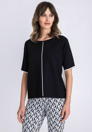 Vrouw draagt een zwart kortärmelig shirt met een witte verticale streep en zwart-witte broeken met geometrisch patroon, staand tegen een effen achtergrond.