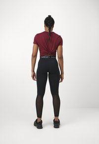 Tumma viininpunainen solmittu T-paita ja mustat legginsit, joissa on verkkomaiset paneelit, yhdistettynä mustiin kenkiin. Legginsit ovat paksulla vyötärönauhalla, jossa on logo.