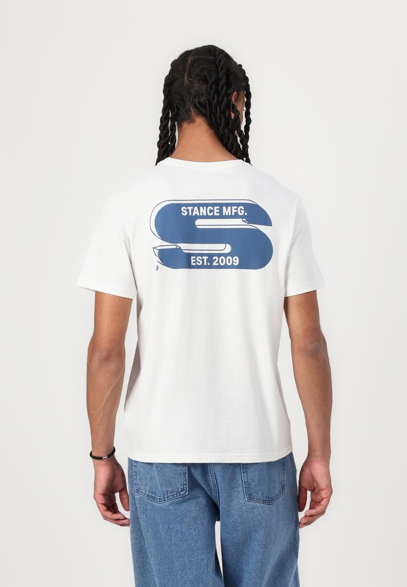 Vit bomull t-shirt med en blå grafik på ryggen, som visar "STANCE MFG." och "ETABL. 2009." Kortärmad och avslappnad passform.