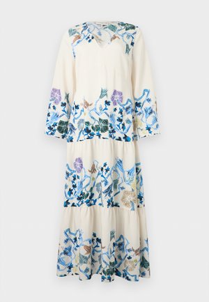 Femme portant une longue robe blanche avec broderies florales bleues, violettes et beiges, manches évasées et talons blancs pointus, debout sur fond blanc.