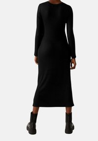 Robe noire à manches longues côtelées avec une silhouette ajustée, longueur mi-mollet, col rond, portée avec des bottines noires épaisses à la cheville.