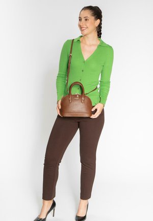 Bruine leren handtas met een gestructureerde afwerking, ronde vorm, dubbele bovenhandvaten en een afneembare schouderband. Model draagt een groene cardigan en een bruine broek.