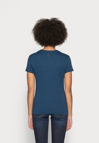 Person med lockigt hår iförd en åtsittande marinblå kortärmad skjorta och mörka jeans, sedd bakifrån mot en enkel bakgrund.