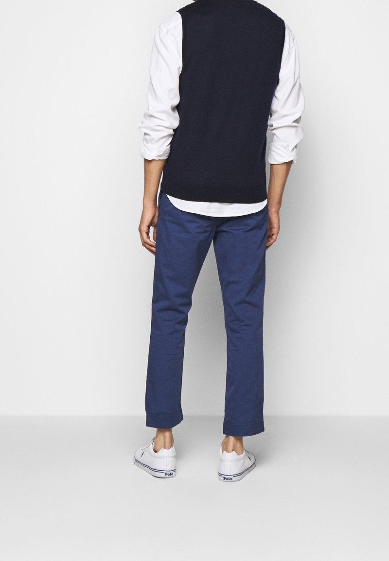 Homme debout de dos, portant une chemise blanche avec manches retroussées, un pull sans manches noir, un pantalon bleu et des baskets blanches sur fond blanc.