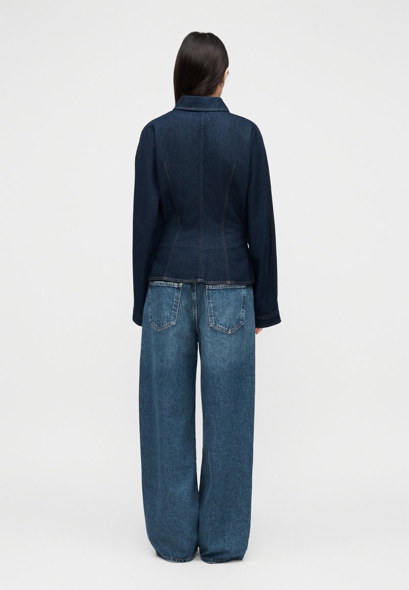 Veste en denim foncé avec une coupe ajustée, dotée d'un col et de surpiqûres contrastantes. Associée à un jean bleu à jambes larges.