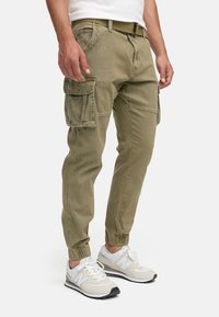 Uomo che indossa pantaloni cargo verde oliva con polsini elasticizzati e una cintura beige, abbinati a scarpe da ginnastica New Balance bianche e grigie.
