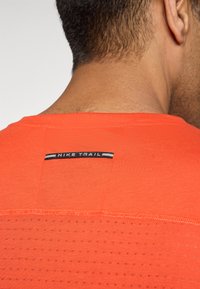 Nærbillede af en persons hals og øvre ryg iført en orange Nike Trail t-shirt med et sort og grå logo mærke ved kraven.