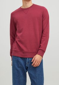 Felpa bordeaux a girocollo con maniche lunghe, caratterizzata da una finitura texturizzata e polsini a costine, abbinata a jeans in denim blu.
