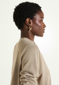 Top de cuello redondo y mangas largas en tejido de punto metálico dorado, con un ajuste relajado. La modelo lleva pendientes de aro dorados. Vista lateral.