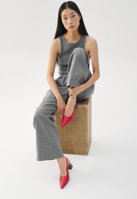 Red slingback heels with a pointed toe and block heel, paired with a gray sleeveless top and wide-leg pants, seated on a wooden cube.