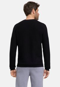 Maglione nero in texture a coste con maniche lunghe, scollo rotondo e orlo aderente. Indossato con pantaloni grigi, che mostrano un design semplice e strutturato.