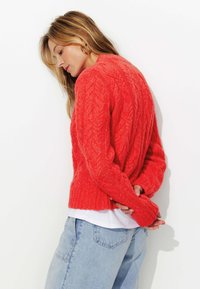 Pull en tricot rouge texturé avec un design court et un ourlet côtelé, associé à un jean à jambe large bleu clair et une chemise blanche en dessous.