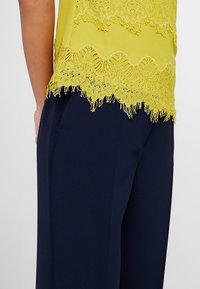 Haut jaune avec une dentelle raffinée et des franges, associé à un pantalon bleu marine foncé. Le matériau semble lisse et léger.