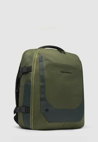 Mochila verde con un tejido texturizado, forma rectangular y acabado mate. Presenta detalles en negro y correas ajustables. Cierres de cremallera visibles.