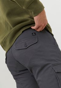 Graue Cargohosen mit seitlichen Taschen, geknöpften Klappen und einem Etikett an der Hintertasche. Getragen mit einem olivgrünen Sweatshirt.