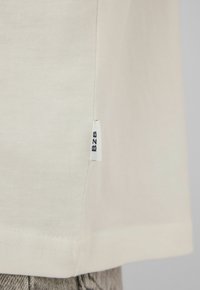 Tissu de couleur crème avec une petite étiquette blanche portant la mention « B2B » cousue dans la couture latérale au-dessus d'un pantalon gris texturé.
