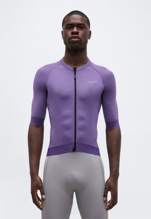 Άνδρας αθλητής με στενό μωβ ποδηλατικό μπλουζάκι με φερμουάρ μπροστά και γκρι επενδυμένο ποδηλατικό σορτς, στέκεται μπροστά σε λευκό απλό φόντο.