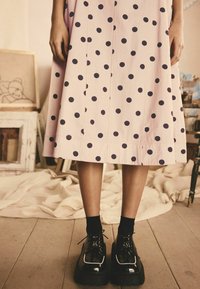 Falda midi de lunares rosas con grandes lunares azul oscuro, que presenta un diseño plisado. Zapatos de plataforma negros con acabado brillante y cordones.