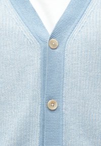 Svetlo modra pletena jakna z dvema bež gumbi, zavezana čez belo notranjo srajco, ki prikazuje rebrasto teksturo in ovratnik v obliki črke V.