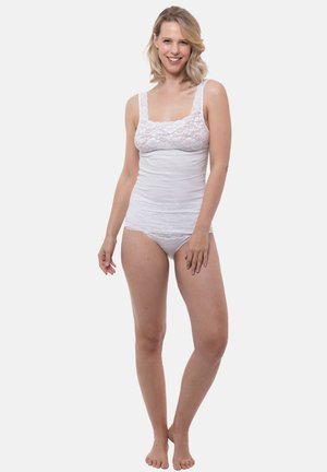 Lächende Frau, barfuß stehend, trägt weiße Spitzentop und passende Unterwäsche vor schlichter Hintergrund.