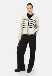 Maglione crema con righe orizzontali nere, colletto a imbuto e dettaglio di fiocco davanti. Abbinato a pantaloni neri larghi e stivali neri chunky.