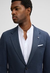 Granatowa marynarka o fakturze z klapą z wycięciem, w połączeniu z białą koszulą z kołnierzykiem typu mandarynka. Posiada zapięcie na guziki oraz ozdobną poszetkę.