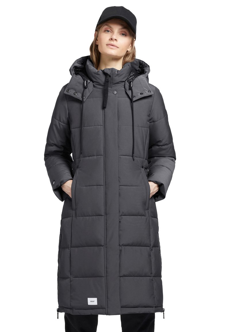 Langer grauer Puffer-Mantel mit Kapuze, ausgestattet mit gesteppten Abschnitten, Kordelzügen, Druckknöpfen und seitlichen Taschen. Schwarze Akzente sind vorhanden.
